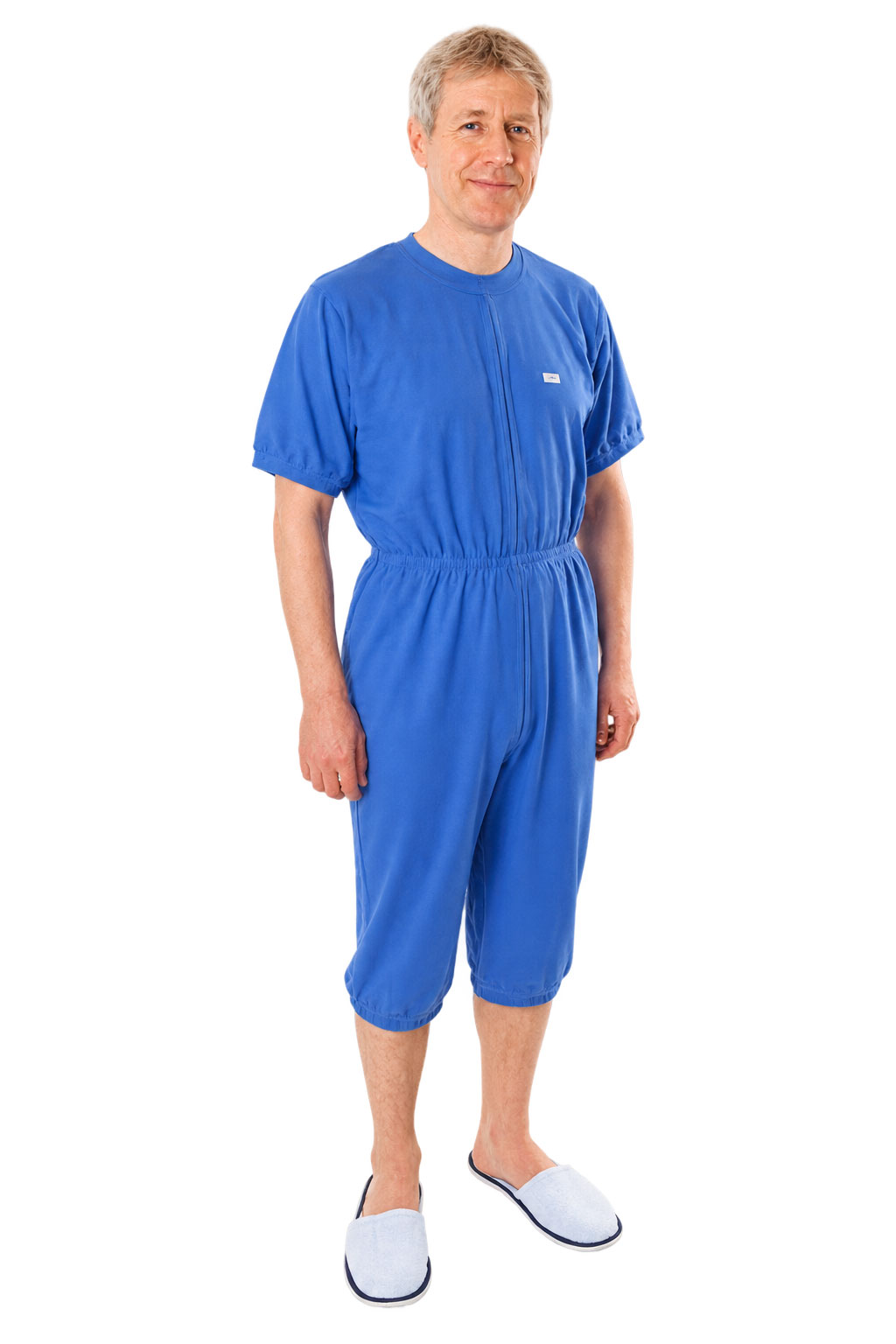 Ein Mann steht aufrecht und trägt einen blauen, kurzärmligen Ganzanzug mit elastischem Bund. Er hat hellblaue Hausschuhe an und lächelt freundlich, vor einem weißen Hintergrund.