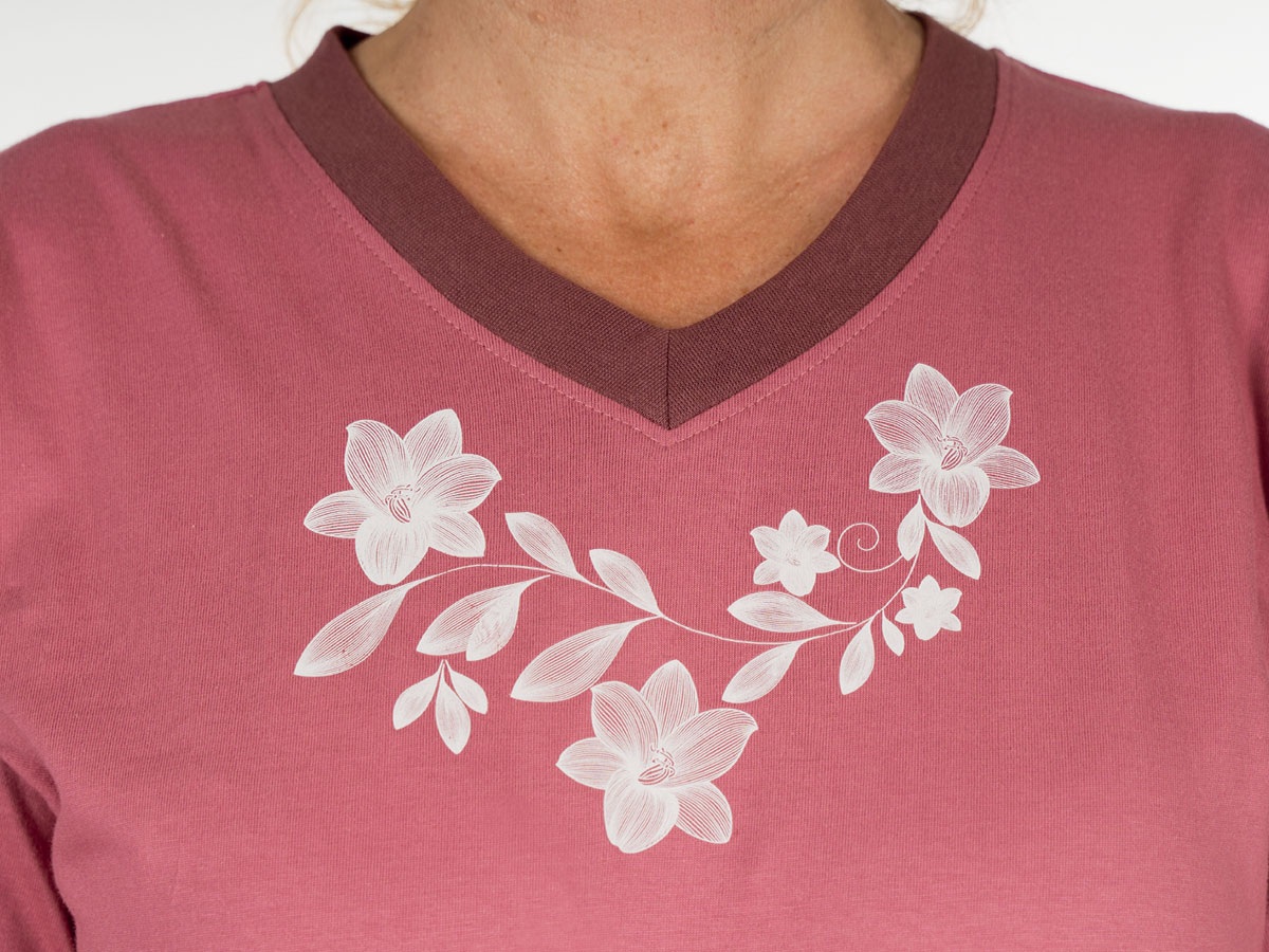 Rosa T-Shirt mit einem V-Ausschnitt, verziert mit einem weißen floralen Muster. Die Blüten und Blätter sind elegant angeordnet und stehen im Kontrast zum Hintergrund des Shirts.