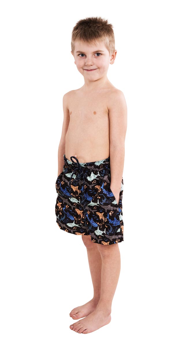 Ein Junge steht barfuß und trägt eine Badehose mit buntem Muster von Tieren. Er lächelt leicht und hat die Hände in den Taschen. Der Hintergrund ist weiß.