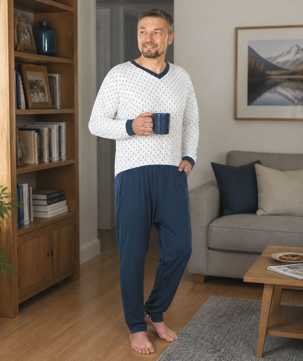 Ein Mann steht in einem Wohnzimmer und trägt einen bequemen Schlafanzug mit einem weißen Oberteil und blauen Unterteilen. Er hält eine Tasse in der Hand und lächelt. Im Hintergrund sind Bücherregale und ein Sofa zu sehen.